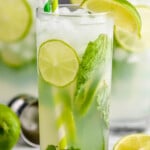 up close view of a high ball glass filled with a mojito cocktail, lime slices, fresh mint, striped green straws, and garnished with lime wedges and fresh mint