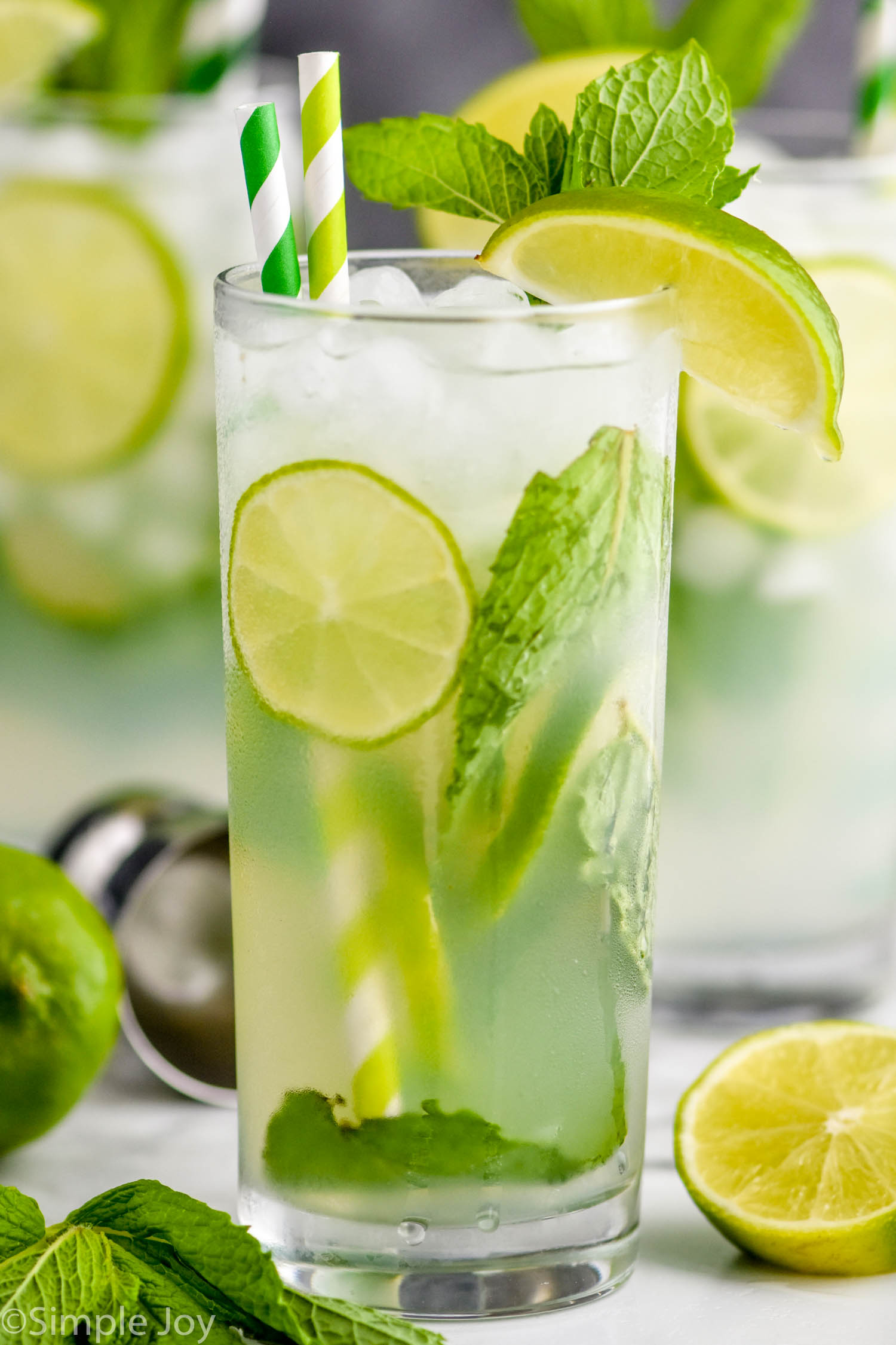 up close view of a high ball glass filled with a mojito cocktail, lime slices, fresh mint, striped green straws, and garnished with lime wedges and fresh mint