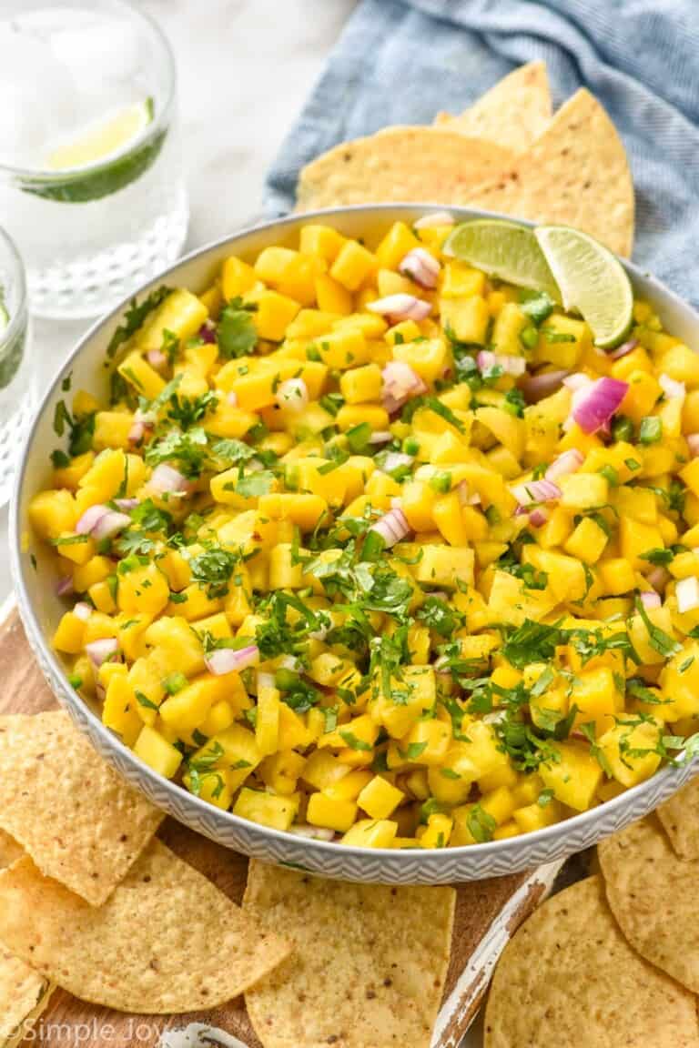 Bowl of Pineapple Mango Salsa garnished with cilantro and lime wedges, with chips and drinks beside