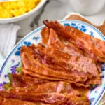 Pinterest graphic for Bacon in The Oven. Text says "how to make bacon in the oven simplejoy.com" Image shows a plate of bacon cooked in the oven with scrambled eggs and fresh strawberries surrounding.