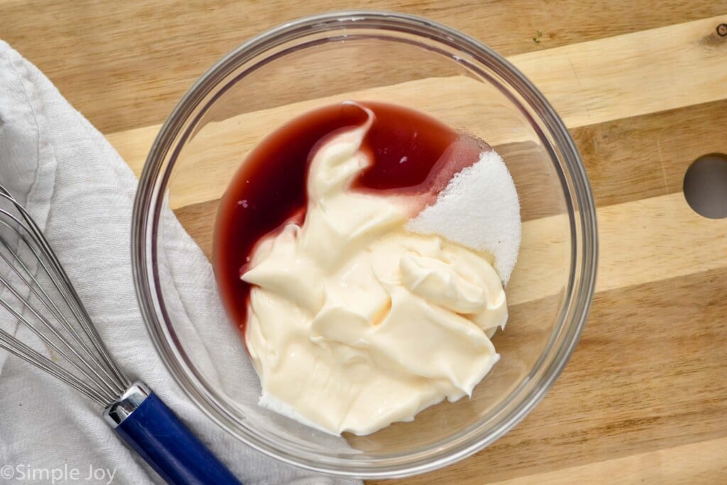 Overhead view of a glass bowl of ingredients for Broccoli Salad dressing recipe. Whisk beside.