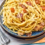 Pinterest graphic for pasta carbonara recipe. Image shows Plate of Pasta Carbonara garnished with parmesan cheese. Forks, glass of red wine, and bread beside. Text says, "pasta carbonara simplejoy.com"