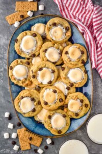 Overhead view of a platter of S'mores Cookies. Glasses of milk, graham crackers, marshmallows, and chocolate chips on counter beside.