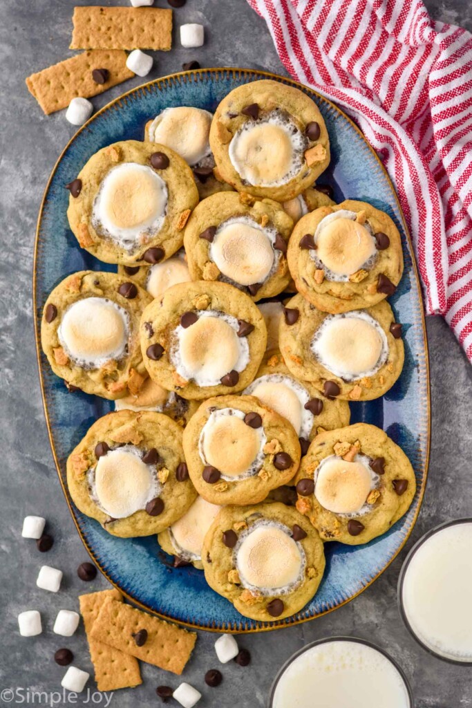 Overhead view of a platter of S'mores Cookies. Glasses of milk, graham crackers, marshmallows, and chocolate chips on counter beside.
