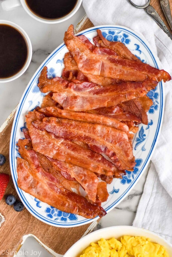 overhead of plate of bacon cooked in the oven. bowl of scrambled eggs, fresh fruit, and two mugs of black coffee sitting beside