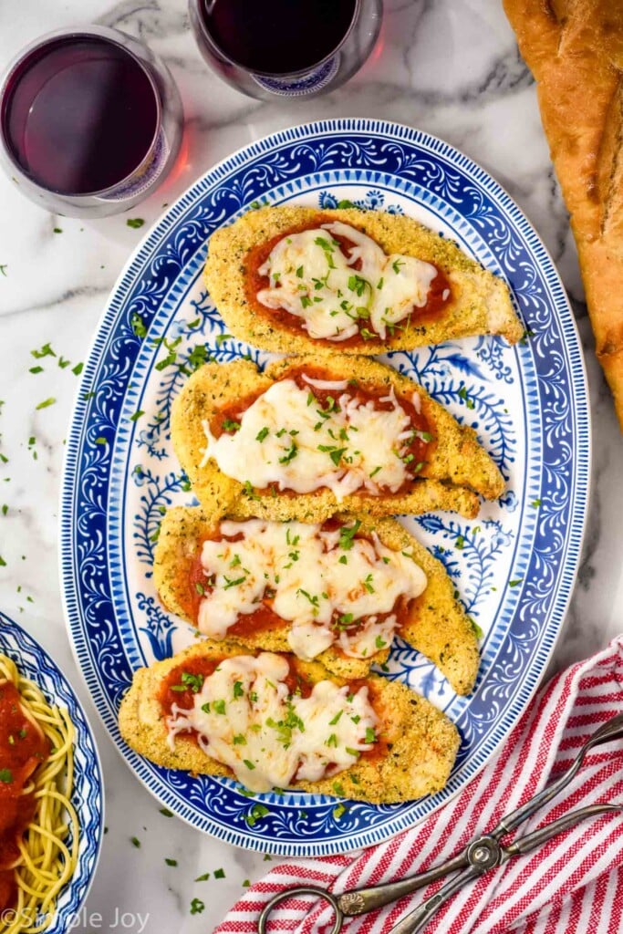 overhead of plate of Air Fryer Chicken Parmesan. Bowl of pasta, glass of red wine, tongs, and bread surrounding plate.