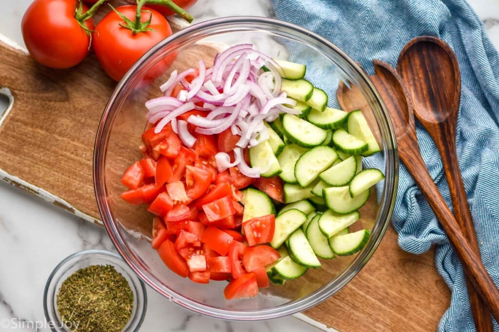 overhead of bowl of Cucumber Tomato Salad ingredients with fresh tomatoes, serving spoons, and bowl of Italian seasoning sitting beside.