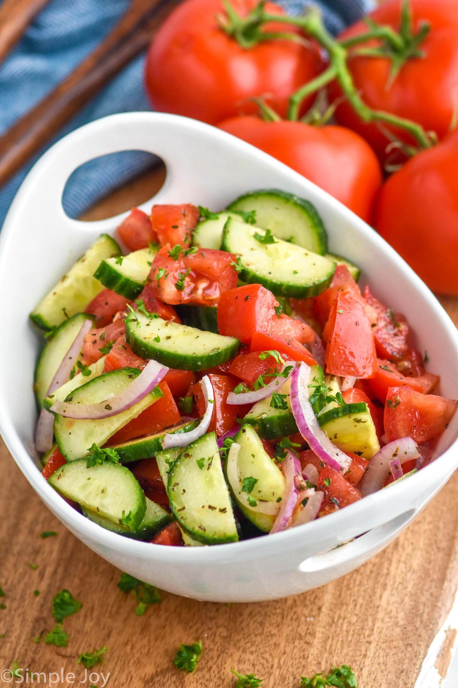 Bowl of Cucumber Tomato Salad sitting on a cutting board. Fresh tomatoes sitting in background