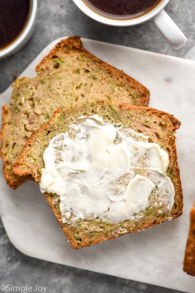 slices of homemade Zucchini Bread on a cutting board. Butter smeared on one slice, mugs of coffee sitting in background