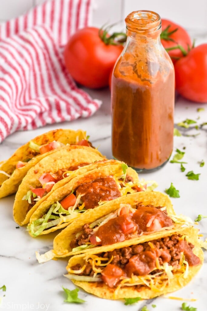 Taco shells with taco ingredients with bottle of homemade taco sauce and tomatoes sitting behind