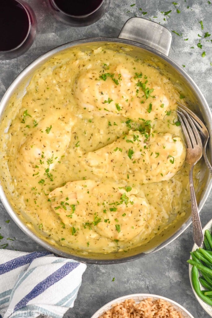 overhead of skillet of Honey Mustard Chicken with two forks. Two glasses of wine, bowl of green beans, and bowl of rice surrounding.