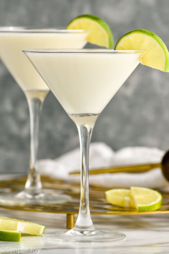 Side view of Key Lime Martini garnished with lime slice. Lime slices and cocktail jigger on counter beside martini glasses.