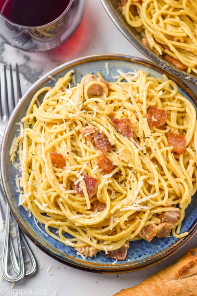 Plate of Pasta Carbonara garnished with parmesan cheese. Forks, glass of red wine, and bread beside.