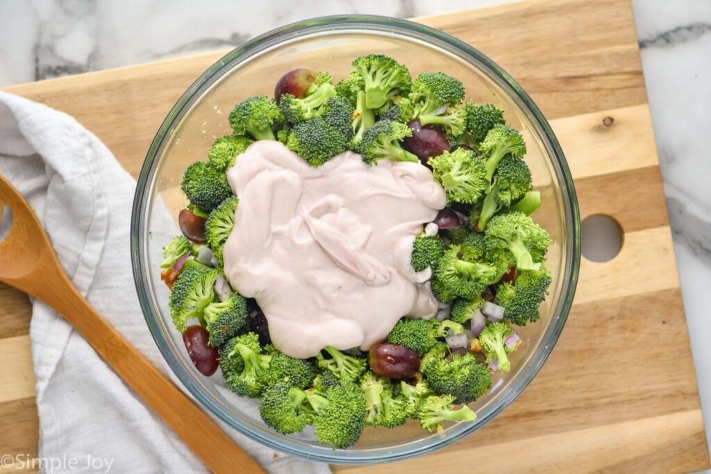 Overhead view of mixing bowl of ingredients and dressing for Broccoli Salad recipe with wooden spoon beside.
