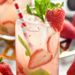 glass of Strawberry Mojito with ice, fresh strawberry and mint garnish, and a straw. Fresh strawberries and Strawberry Mojito sitting in background. - 10