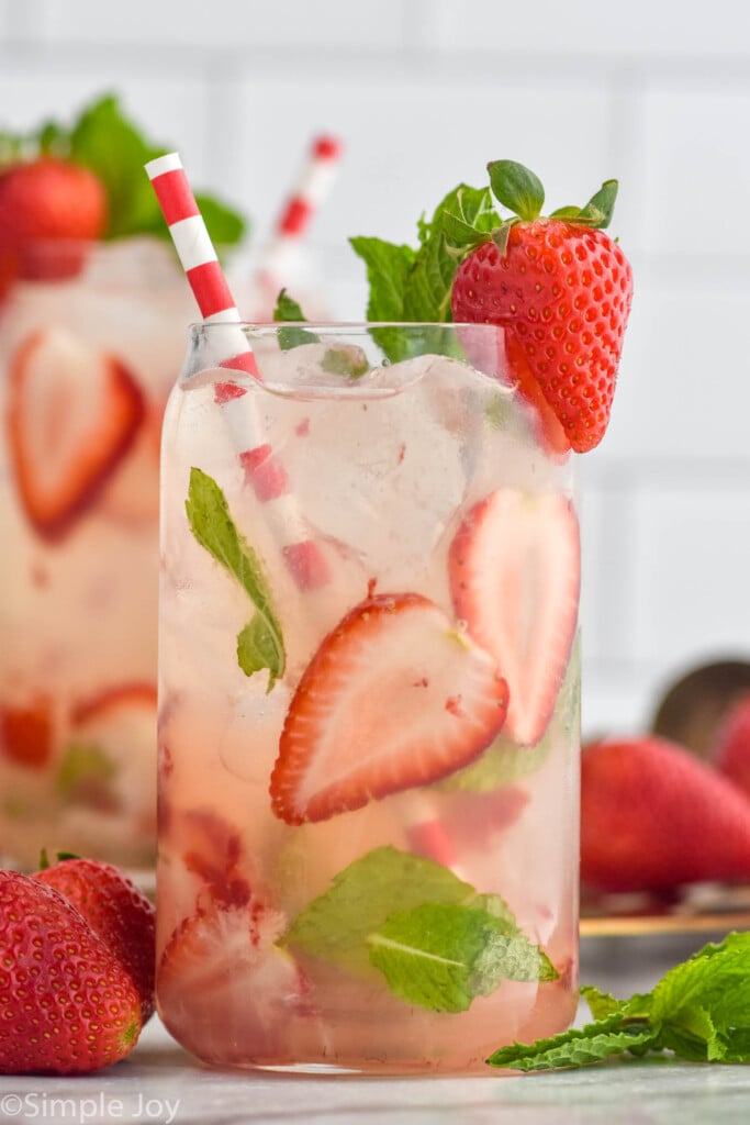 glass of Strawberry Mojito with ice, fresh strawberry and mint garnish, and a straw. Fresh strawberries and Strawberry Mojito sitting in background.