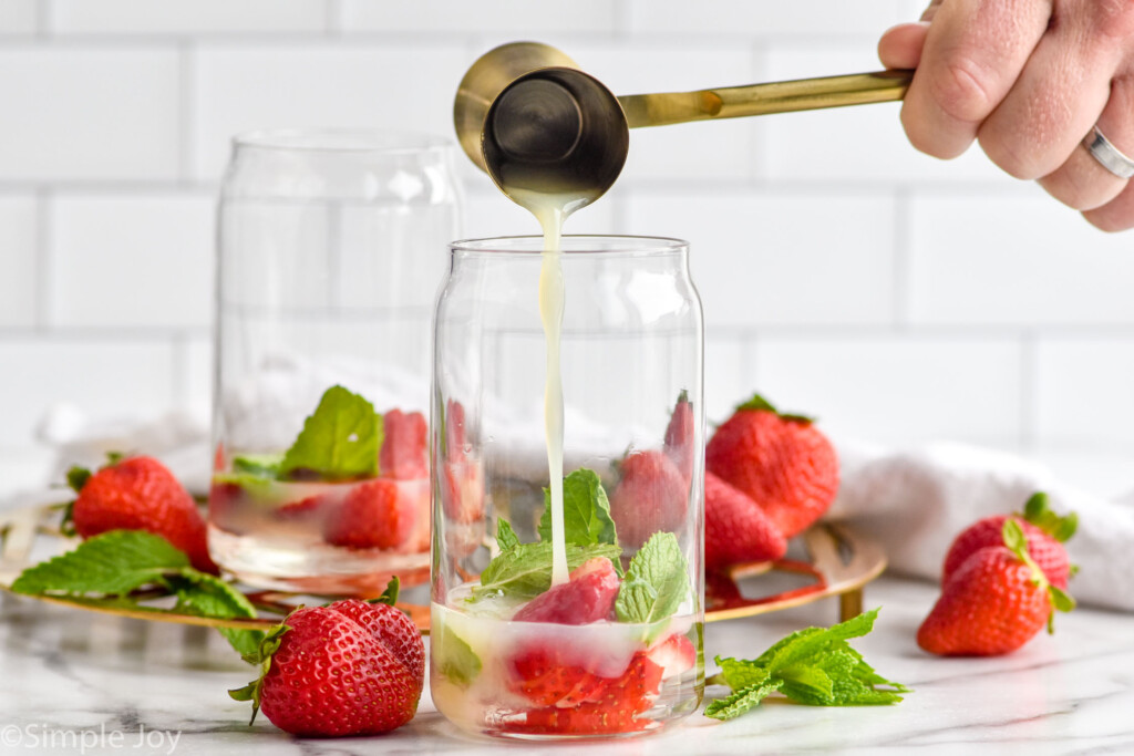 man's hand pouring cocktail jigger of lime juice into a glass of mint leaves and strawberries to make a Strawberry Mojito cocktail. Fresh strawberries and mint leaves sitting in background.