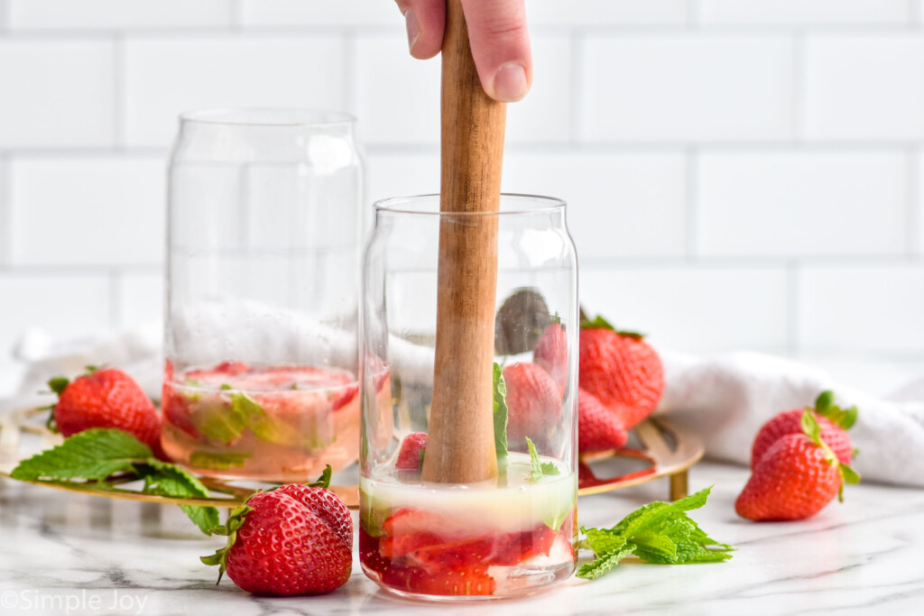 man's hand muddling strawberry mojito ingredients in a glass. Fresh strawberries and mint leaves sitting in background