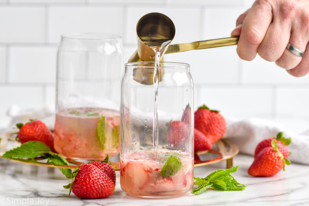 man's hand pouring cocktail jigger of rum into a glass of mint leaves and strawberries to make a Strawberry Mojito cocktail. Fresh strawberries and mint leaves sitting in background.