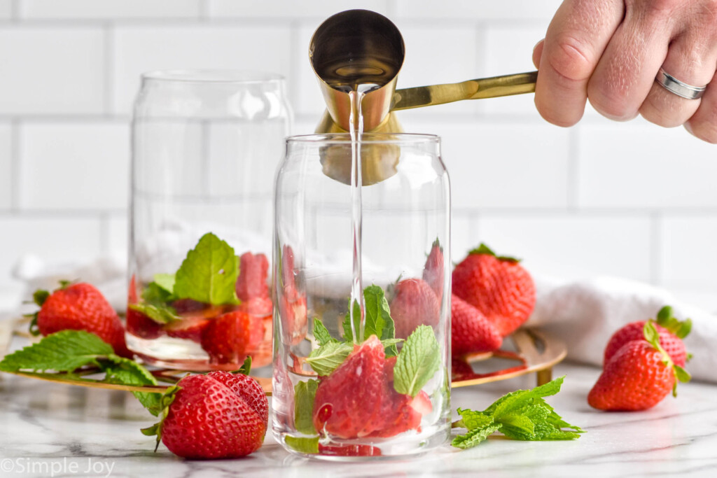 man's hand pouring cocktail jigger of simple syrup into a glass of mint leaves and strawberries to make a Strawberry Mojito cocktail. Fresh strawberries and mint leaves sitting in background.