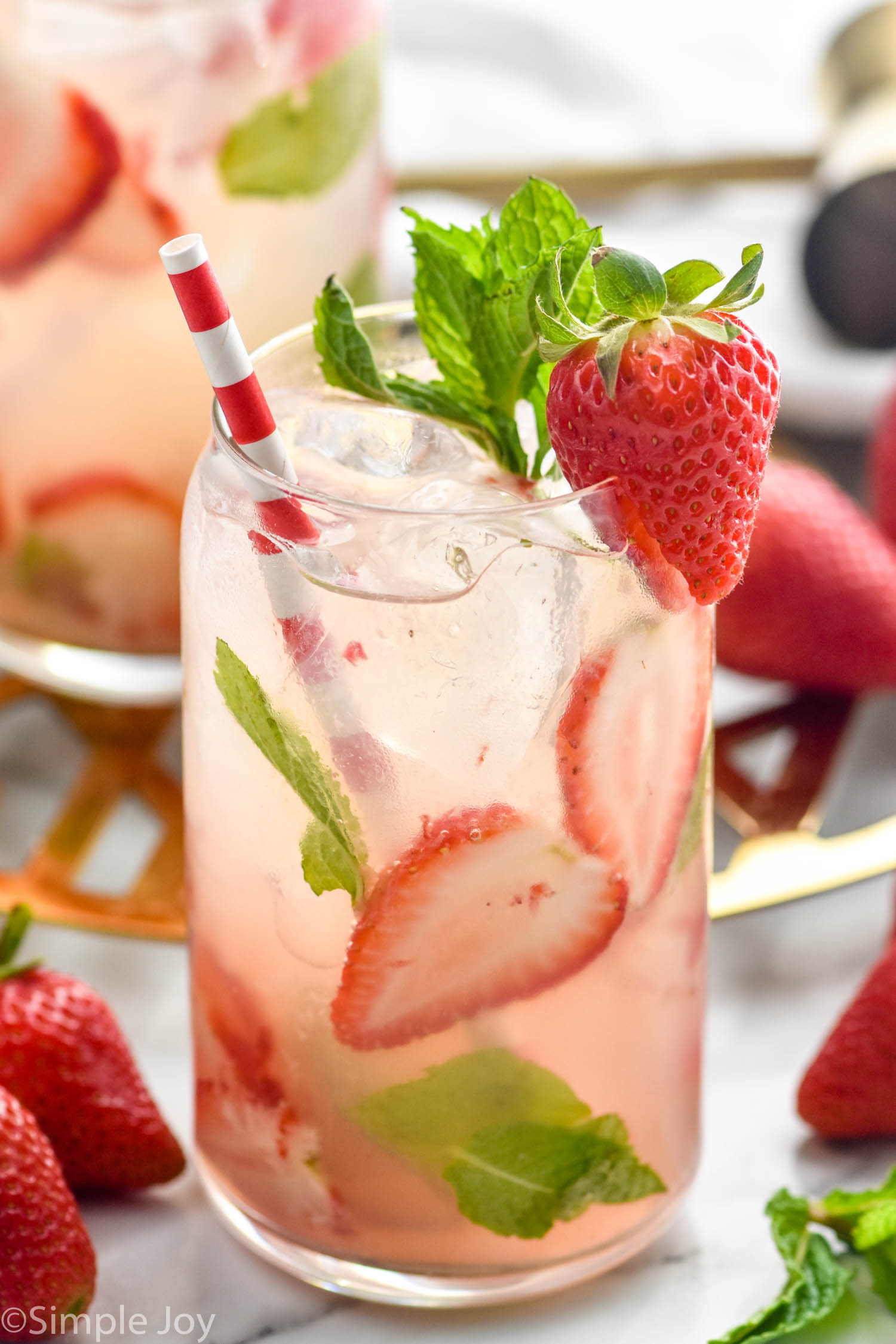 glass of Strawberry Mojito with ice, fresh strawberry and mint garnish, and a straw. Fresh strawberries and Strawberry Mojito sitting in background.