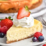 Plate of piece of Easy Cheesecake Recipe stopped with homemade whipped cream and a strawberry. Fresh raspberries and blueberries sitting beside. Cheesecake topped with berries sitting in background - 15