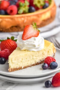 Plate of piece of Easy Cheesecake Recipe stopped with homemade whipped cream and a strawberry. Fresh raspberries and blueberries sitting beside. Cheesecake topped with berries sitting in background