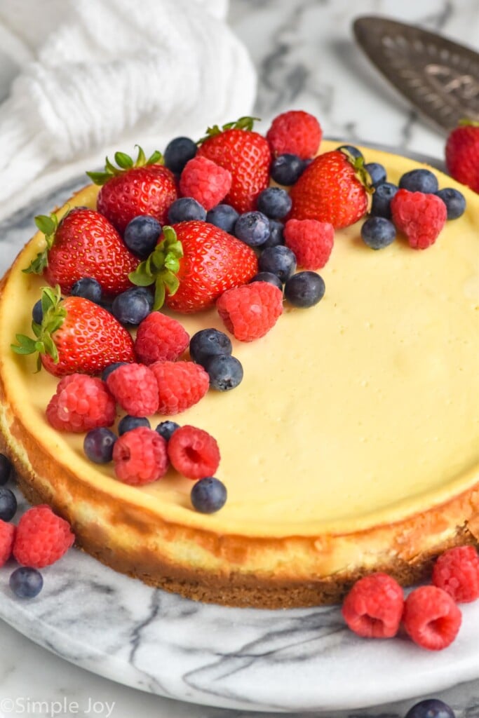 Easy Cheesecake Recipe topped with fresh strawberries, blueberries, and raspberries. Fresh berries and serving spatula sitting beside.