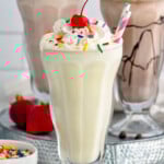 Glass of vanilla milkshake topped with whipped cream, sprinkles, and a cherry. Glasses of strawberry milkshake, chocolate milkshake, and fresh strawberries sitting in background. - 10