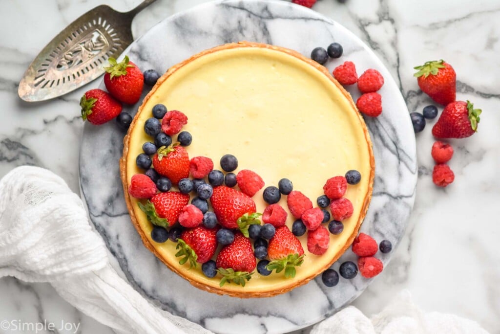 overhead of Easy Cheesecake Recipe topped with fresh strawberries, blueberries, and raspberries. Serving spatula and fresh berries sitting beside.