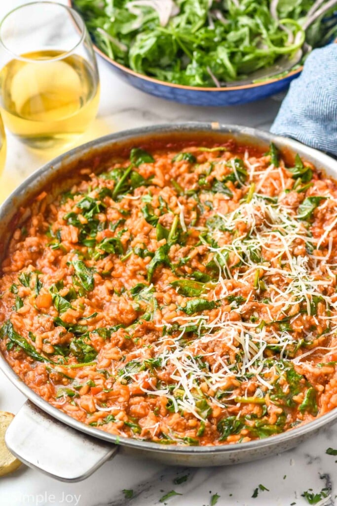 skillet of risotto with spinach and shredded parmesan cheese. Bowl of salad and glass of white wine sitting in background
