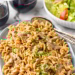 Pinterest graphic for Beef Stroganoff. Text says "the best Beef Stroganoff simplejoy.com" Image shows a serving bowl of Beef Stroganoff with two spoons for serving, bowl of lettuce salad and two glasses of red wine sitting in background.