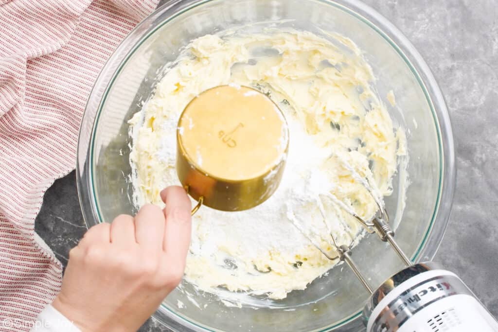 a hand dumping a cup of powdered sugar into a mixing bowl with creamed together cream cheese and butter