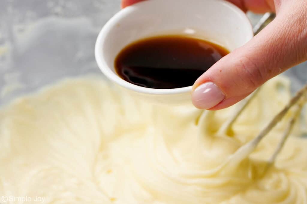 a hand pouring a small amount of vanilla extract into a bowl of cream cheese frosting