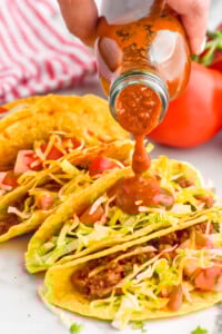 Women's hand pouring bottle of homemade taco sauce over taco shells filled with taco meat, shredded lettuce, shredded cheese, and diced tomatoes. - 8