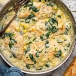 Overhead of skillet of spinach artichoke chicken with two forks. Glass of white wine and baguette sitting beside