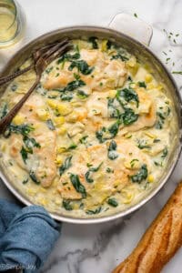 Overhead of skillet of spinach artichoke chicken with two forks. Glass of white wine and baguette sitting beside