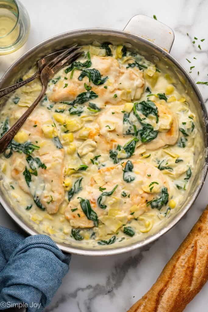 Overhead of skillet of spinach artichoke chicken with two forks. Glass of white wine and baguette sitting beside