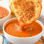 Bowl of Tomato Soup garnished with parsley with half of grilled cheese sandwich being dipped in it. Another bowl of Tomato Soup and spoons beside. - 10