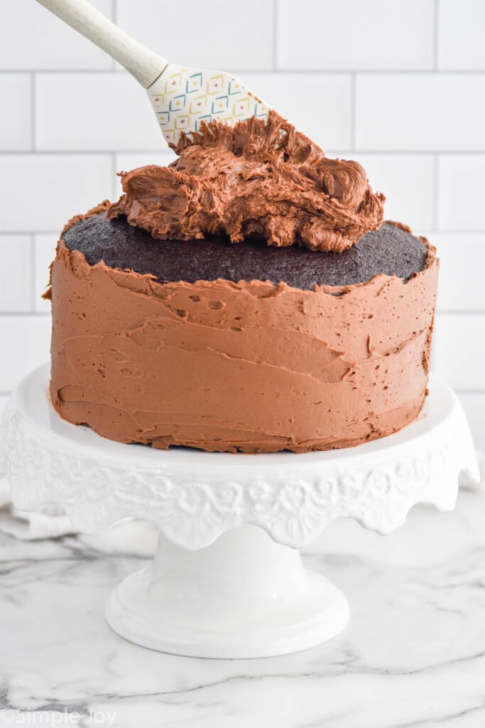 Chocolate Cake sitting on cake stand with sides frosted, dollop of frosting and spatula on top of the cake