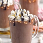 mug of homemade Hot Chocolate topped with marshmallows and chocolate sauce. Two mugs of Hot Chocolate sitting in background, marshmallows sitting beside - 8