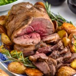platter of Boneless Leg of Lamb, potatoes, and rosemary sprigs. Two glasses of red wine and bowl of salad sitting in background - 13