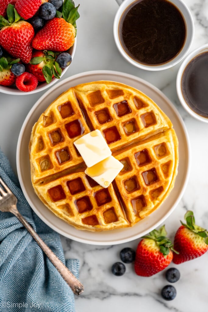 overhead of waffles on a plate with two pieces of butter, syrup, a bowl of fruit, and some coffee