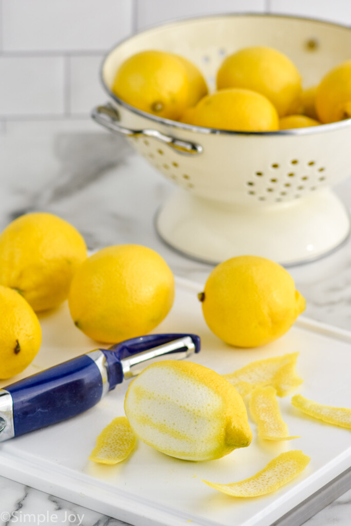 Lemons on cutting board with peeler next to peeled lemon. Colander of more lemons beside for Limoncello recipe.