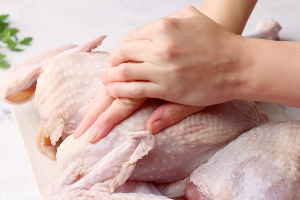 two hands pressing down on a raw turkey to break the breast bone