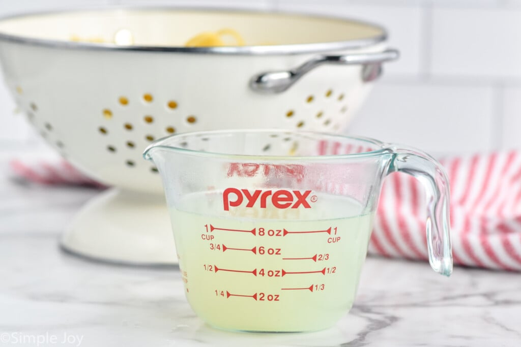 glass measuring cup of reserved pasta water with colander of cooked pasta sitting in background