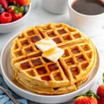 two waffles stacked on a white plate with two pieces of butter and syrup, a bowl of fruit and coffee in the background