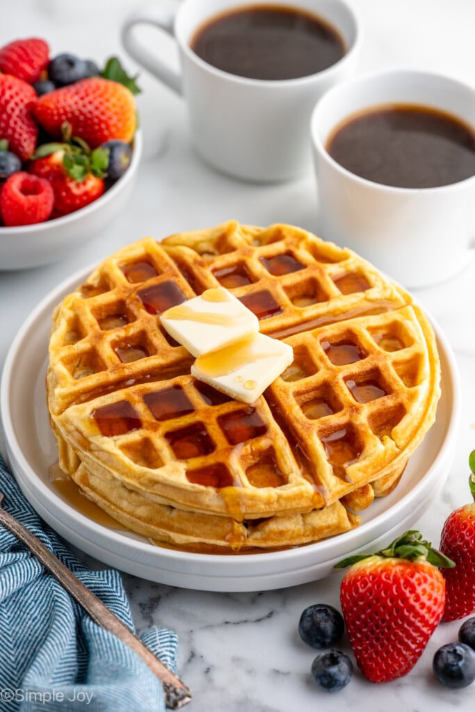 two waffles stacked on a white plate with two pieces of butter and syrup, a bowl of fruit and coffee in the background