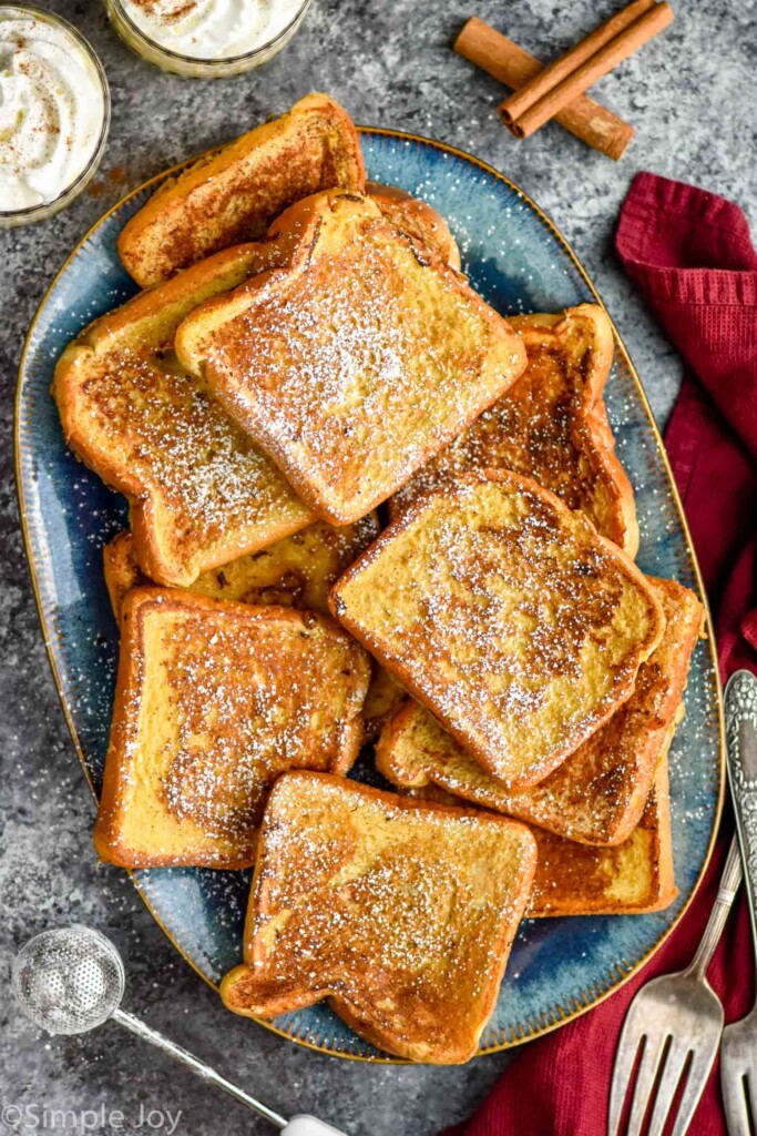 overhead of plate of Eggnog French Toast topped with powdered sugar. Two glasses of eggnog and cinnamon sticks sitting in background