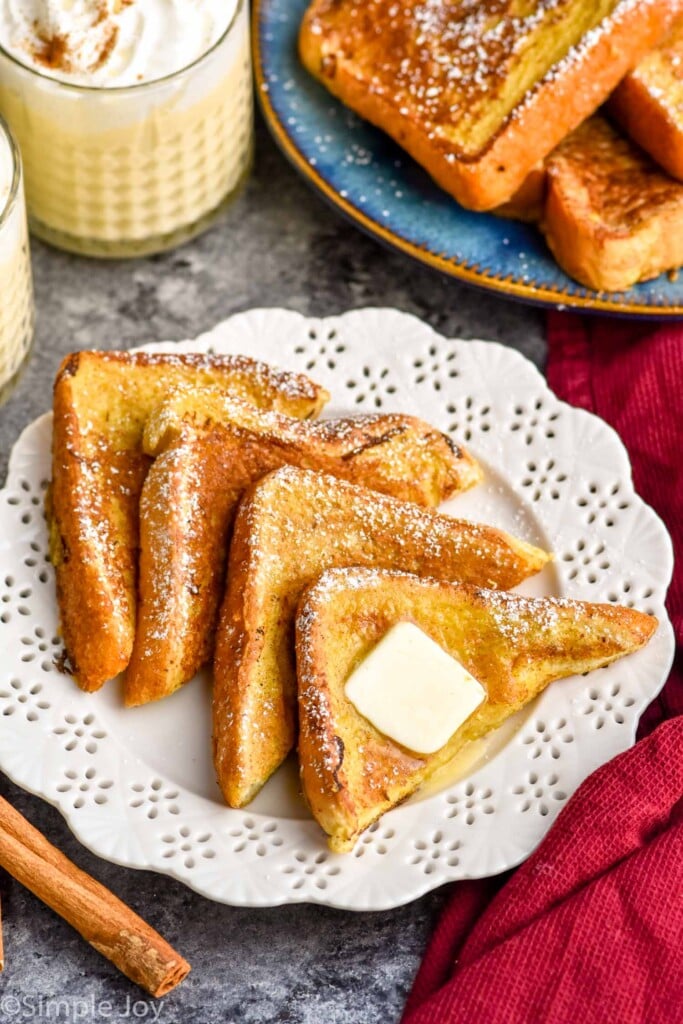 plate of Eggnog French Toast topped with powdered sugar and butter. Two glasses of eggnog and plate of Eggnog French Toast sitting behind